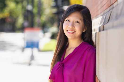Participant Lena - person with long dark hair in a pink blouse leaning against a brick building