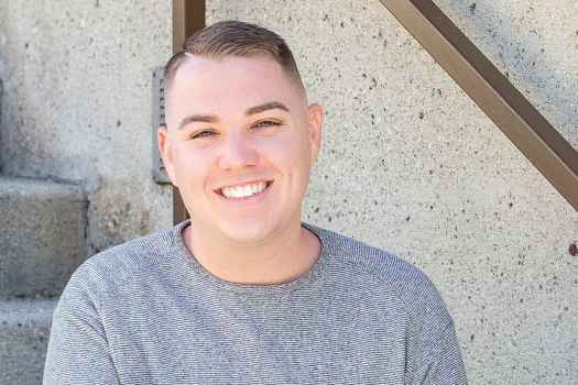 Participant Kyle - person with short brown hair in a grey sweater smiling and sitting on concrete steps