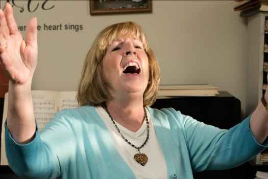 Participant Lisa - person with blond hair in a blue sweater singing in front of a piano