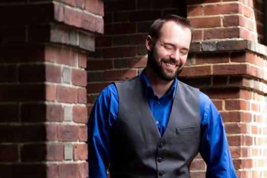 Participant Nathaniel - person with short dark hair wearing a blue shirt leaning against a brick pillar