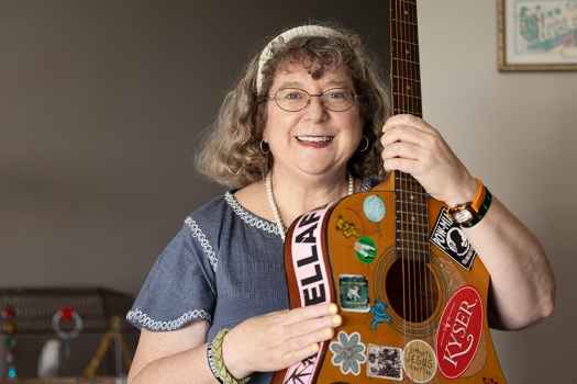 Participant Mary - person with wavy grey hair smiling with a guitar