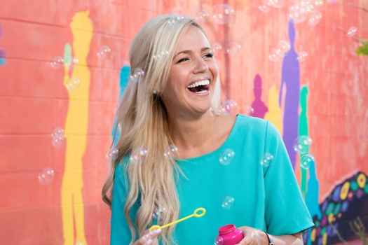 Participant Meg - person with long blond hair laughing in front of a colorful painted wall holding a bubble wand and surrounded by bubbles