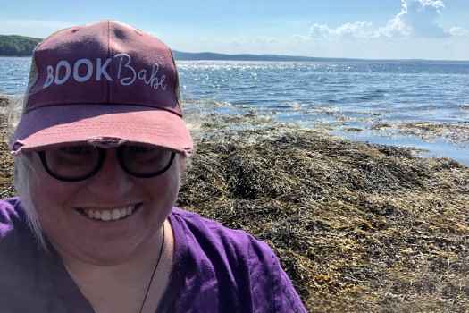 Participant Vanessa - person with hair tied back, smiling in front of a beach, wearing glasses and a hat that says book babe