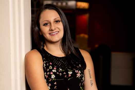 Participant Jackie - person with long dark hair smiling and leaning against a wall