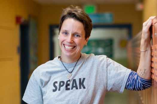 Participant Jen - person with short auburn hair wearing a grey shirt that says speak leaning against orange lockers in a hallway