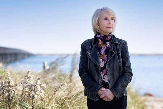 Participant Joanne - person with short white hair wearing a colorful patterned scarf standing in front of a beach with a bridge