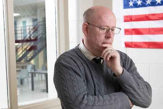 Participant John - person with glasses wearing a grey sweater in front of an office window and an American flag