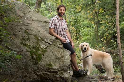 Participant Jeff - person with short and curly brown hair leaning against a large rock holding a leash for a golden retriever