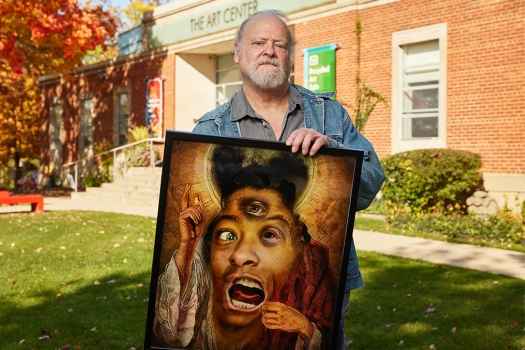 Participant David - person with white hair wearing blue denim outside surrounded by trees with autumn leaves and holding a framed piece of artwork