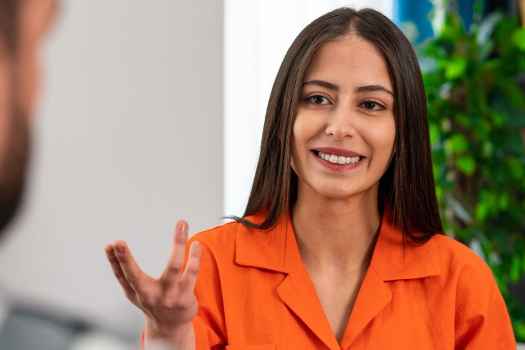 Woman in orange shirt speaking to someone in front of a bookshelf with plants
