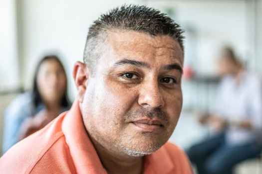 Portrait of a person with short hair wearing a coral shirt. There are other people in the background