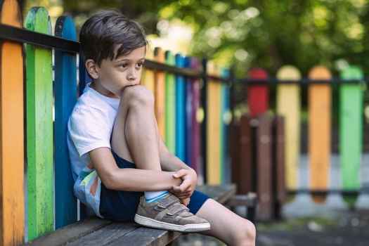 Young child sitting with a knee up on a bench in front of a colored wooded fence