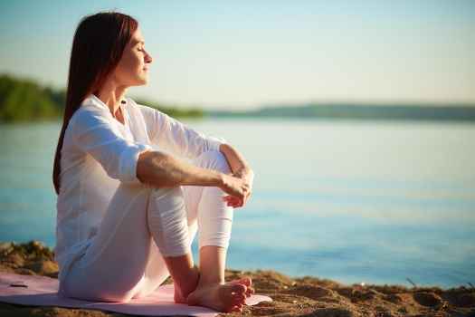 Woman sits by water, eyes closed knees up