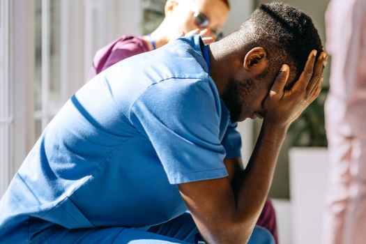 Man in scrubs sits holding his head in his hands