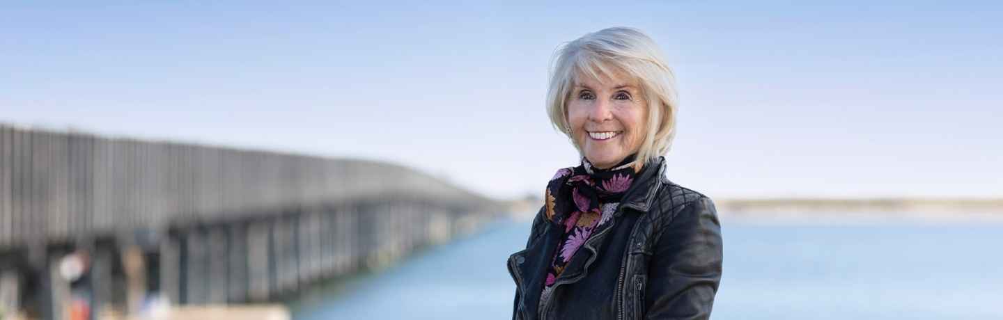 Participant Joanne - person with short white hair wearing a colorful patterned scarf and smiling in front of a beach with a bridge