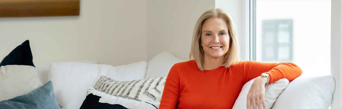 Participant Carrie - person with shoulder-length blond hair wearing an orange shirt and smiling, sitting on a white couch