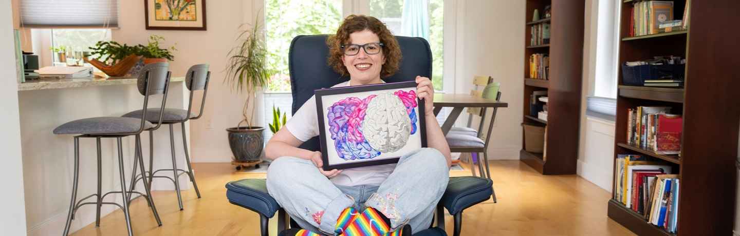 Participant Caitlin - person with short curly hair and glasses smiling, holding a framed illustration