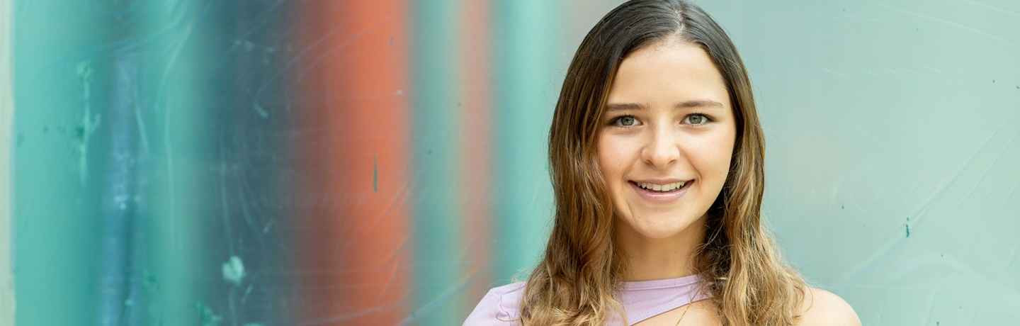 Participant Alejandra - person with brown wavy hair and a purple shirt smiling in front of a bright teal wall
