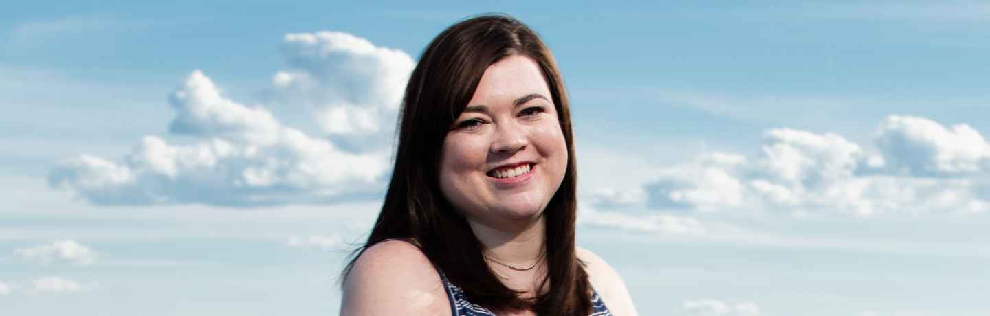 Participant Clare - person with long dark hair smiling in front of a blue sky