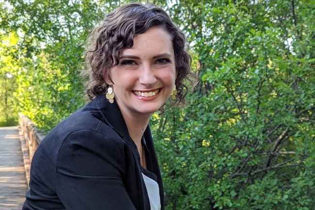 Participant Katie - person in black blazer and blue shirt smiling in front of greenery