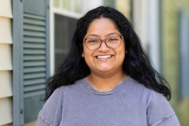 Participant Marina - person with dark hair and glasses wearing a grey t-shirt smiling in front of a building