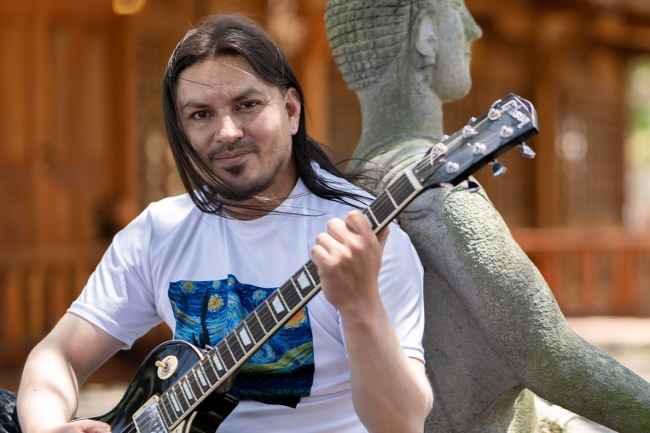 Participant Pablo - person with long dark hair holding a guitar and leaning against a statue