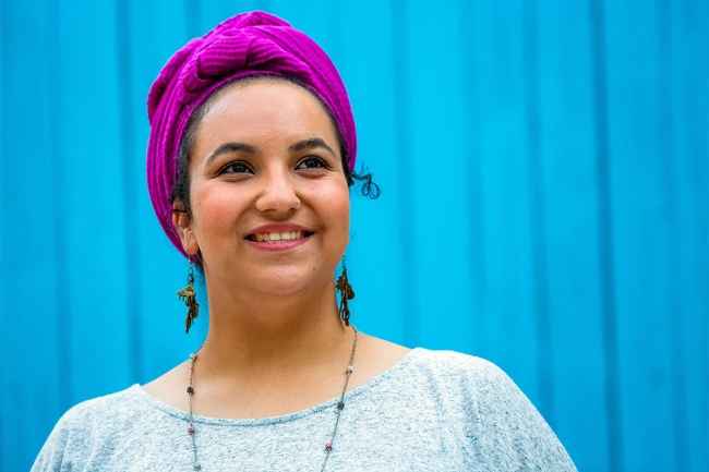 Deconstructing Stigma participant Leah Roni - person in headscarf against bright blue fence