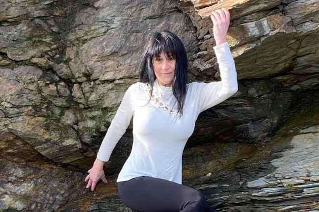 Participant Nique - person with long dark hair wearing a white turtleneck shirt leaning against a large rock