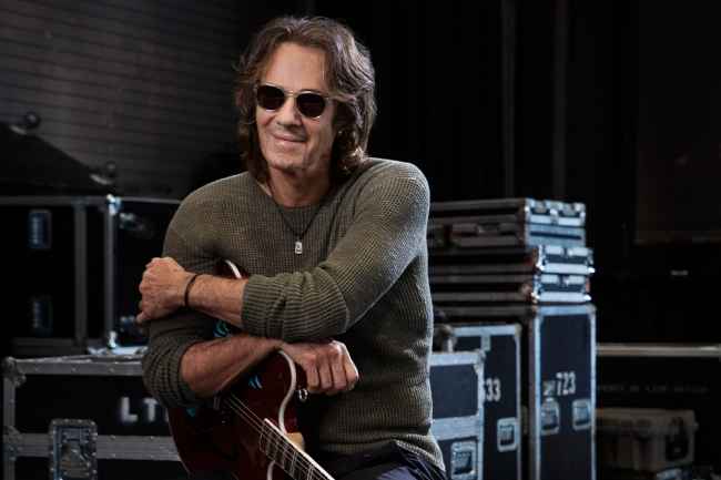 Participant Rick - person with long hair and sunglasses in front of a dark background holding a guitar