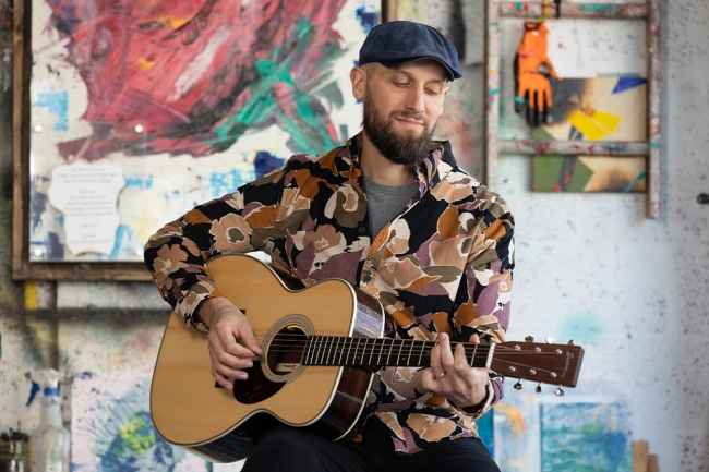 Participant Edmund - person with dark facial hair wearing a dark hat and a colorful patterned shirt playing a guitar