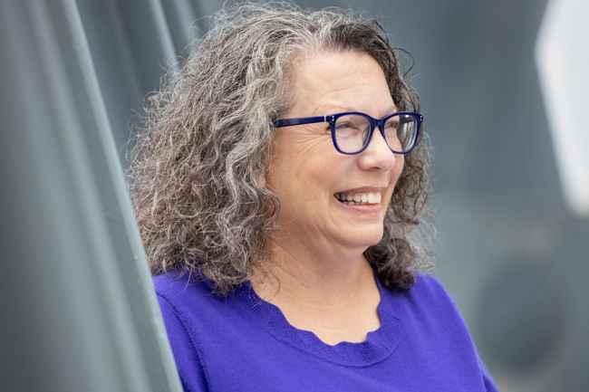 Participant April - person with curly grey hair wearing a purple shirt and smiling in front of a grey background