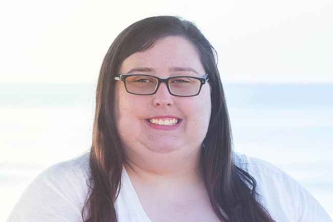 Participant Elizabeth - person with long dark hair and glasses smiling outside in front of a blue sky