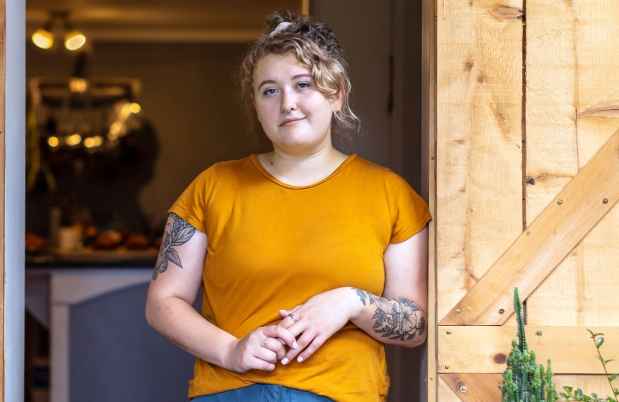 Participant Lexi - person with curly hair in an orange shirt leaning on a wooden door