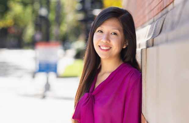 Participant Lena - person with long dark hair in a pink blouse leaning against a brick building