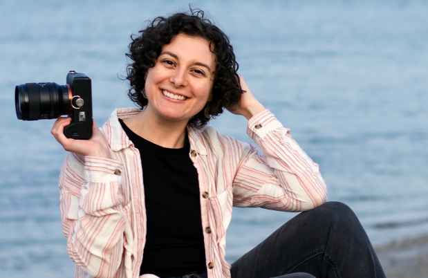 Participant Marena - person smiling holding a camera outside by a beach