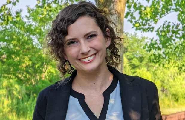 Participant Katie - person in black blazer and blue shirt smiling in front of greenery