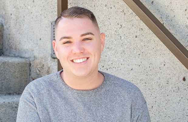 Participant Kyle - person with short brown hair in a grey sweater smiling and sitting on concrete steps