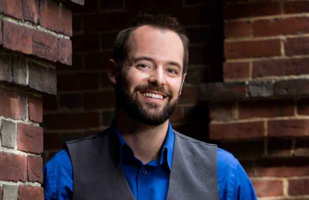 Participant Nathaniel - person with short dark hair wearing a blue shirt leaning against a brick pillar