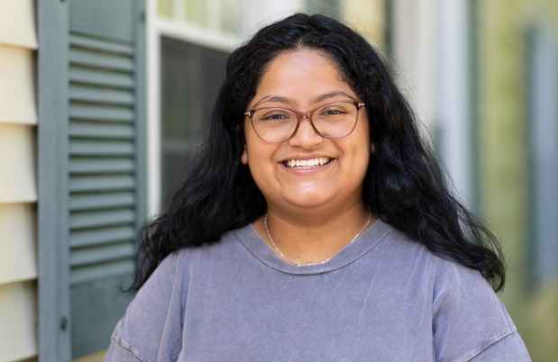 Participant Marina - person with dark hair and glasses wearing a grey t-shirt smiling in front of a building