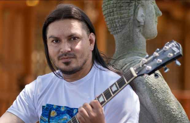 Participant Pablo - person with long dark hair holding a guitar and leaning against a statue