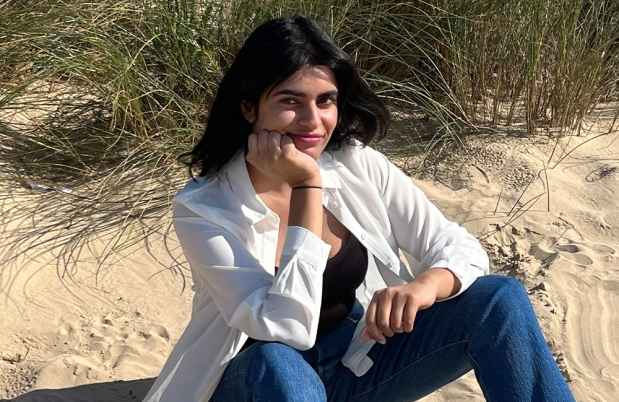 Participant Samina - person wearing jeans and a white coat sitting on sand at a beach