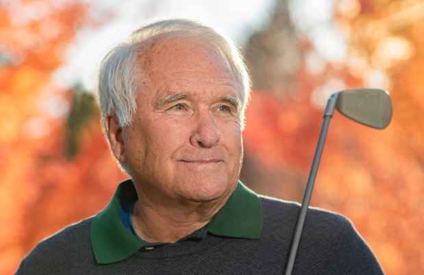 Participant Mike - person with short white hair holding a golf club in front of fall foliage
