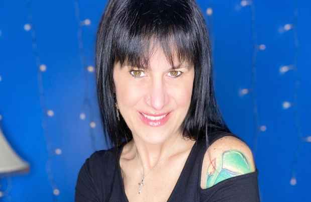 Participant Nique - person with long dark hair smiling with crossed arms in front of a blue wall