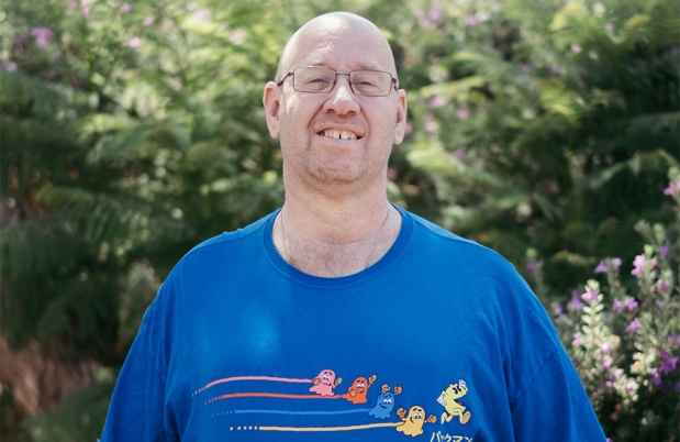 Deconstructing Stigma participant Ori - Person in blue shirt stands outside greenery behind