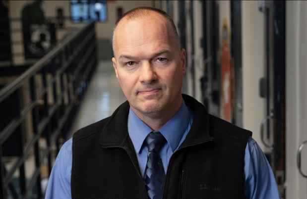 Participant Randall - a person wearing a blue shirt and a black vest in front of a hallway