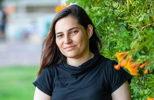 Deconstructing Stigma participant Shelly - Person in black shirt and long hair stands outside with greenery behind