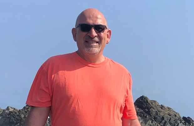 Participant Steve - person wearing sunglasses and a bright coral shirt smiling in front of a body of water with large rocks