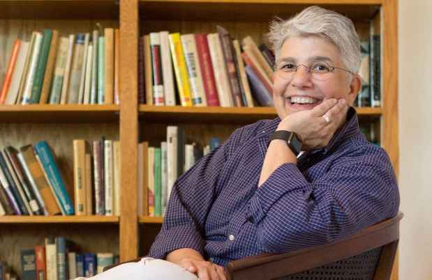 Participant Olga - person with short white hair wearing a blue shirt smiling in front of a bookshelf