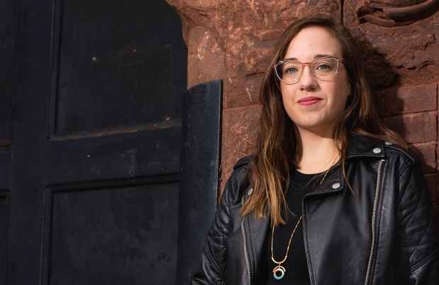 Participant Angi - person with long wavy hair and glasses wearing a black leather jacket and leaning against a stone building