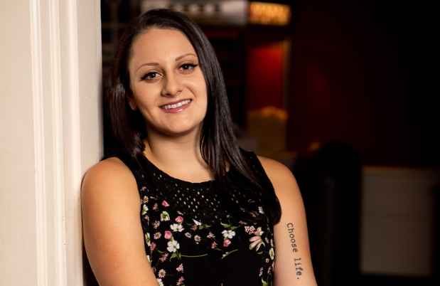 Participant Jackie - person with long dark hair smiling and leaning against a wall